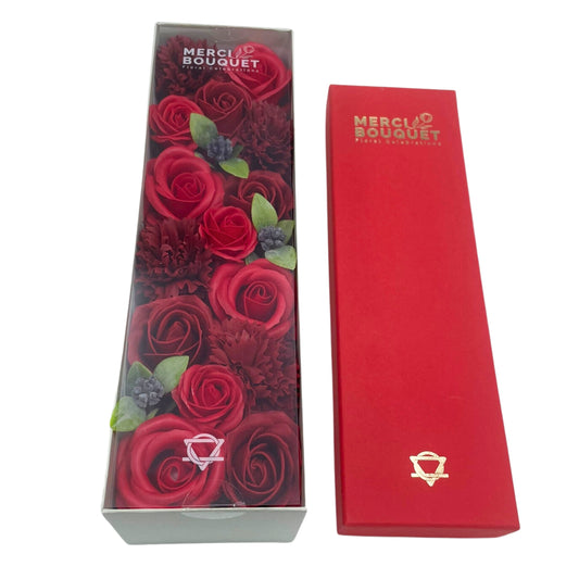 A bouquet of red and burgundy roses and carnations sits inside a clear plastic-topped box; a matching red box sits beside it. Both boxes have a gold logo that reads "MERCI BOUQUET Floral Celebrations".