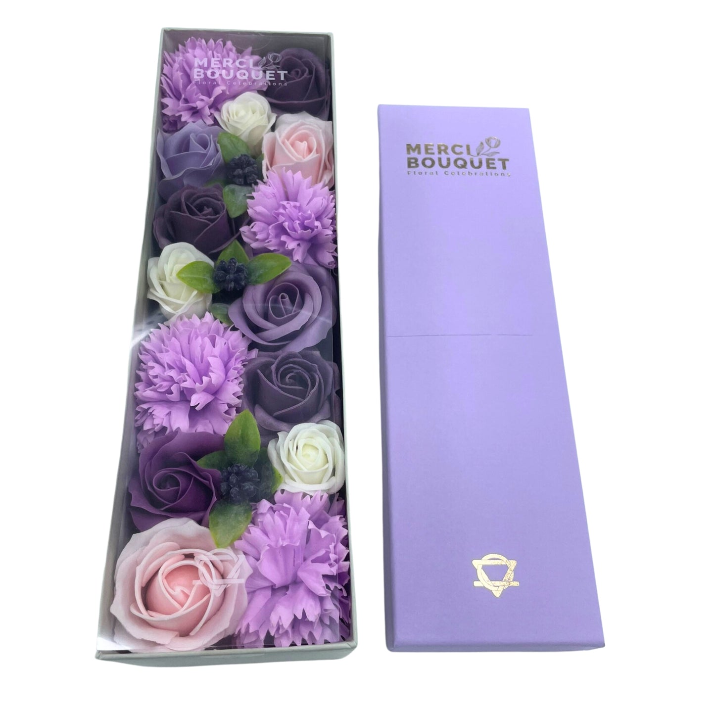 Soap flowers in shades of purple and pink are arranged in a clear box. The box sits next to a lavender-colored gift box. The boxes are labeled "MERCI BOUQUET Floral Celebrations".