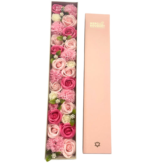 A long box containing a pink and white soap flower arrangement sits next to its matching lid. MERCI BOUQUET Floral Celebrations is printed on both the box and its lid.