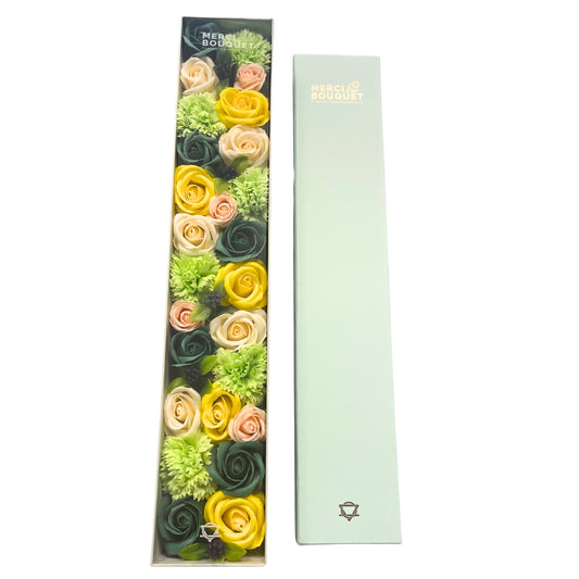 A long, rectangular box containing an arrangement of yellow, peach, and green soap roses and carnations sits beside its light-green outer box. MERCI BOUQUET Floral Celebrations is printed on both.