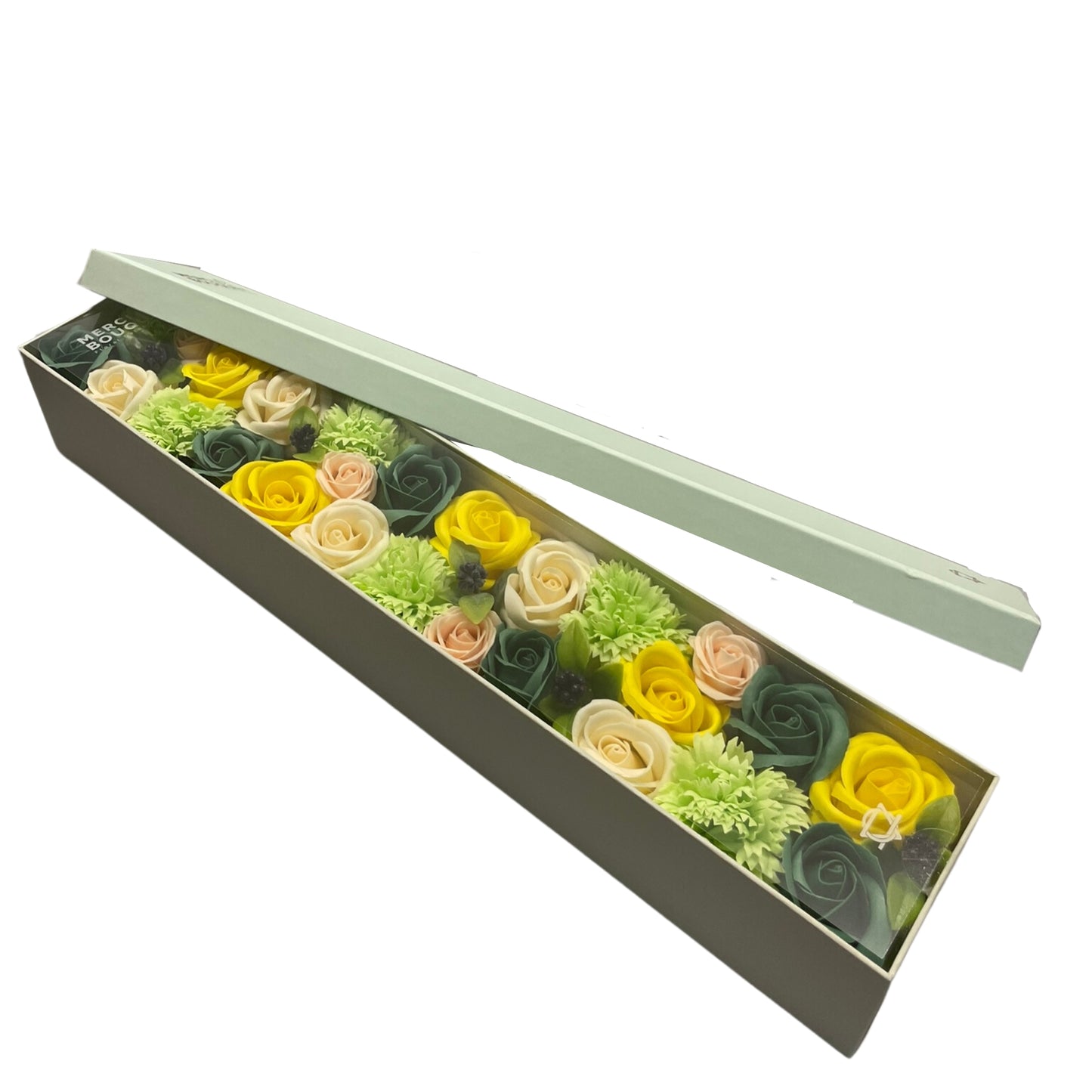 A long pale-green box containing a floral arrangement of yellow, peach, and dark-green soap roses and carnations rests on a white background; its lid is ajar. MERCI BOUQUET is partially visible on the box.