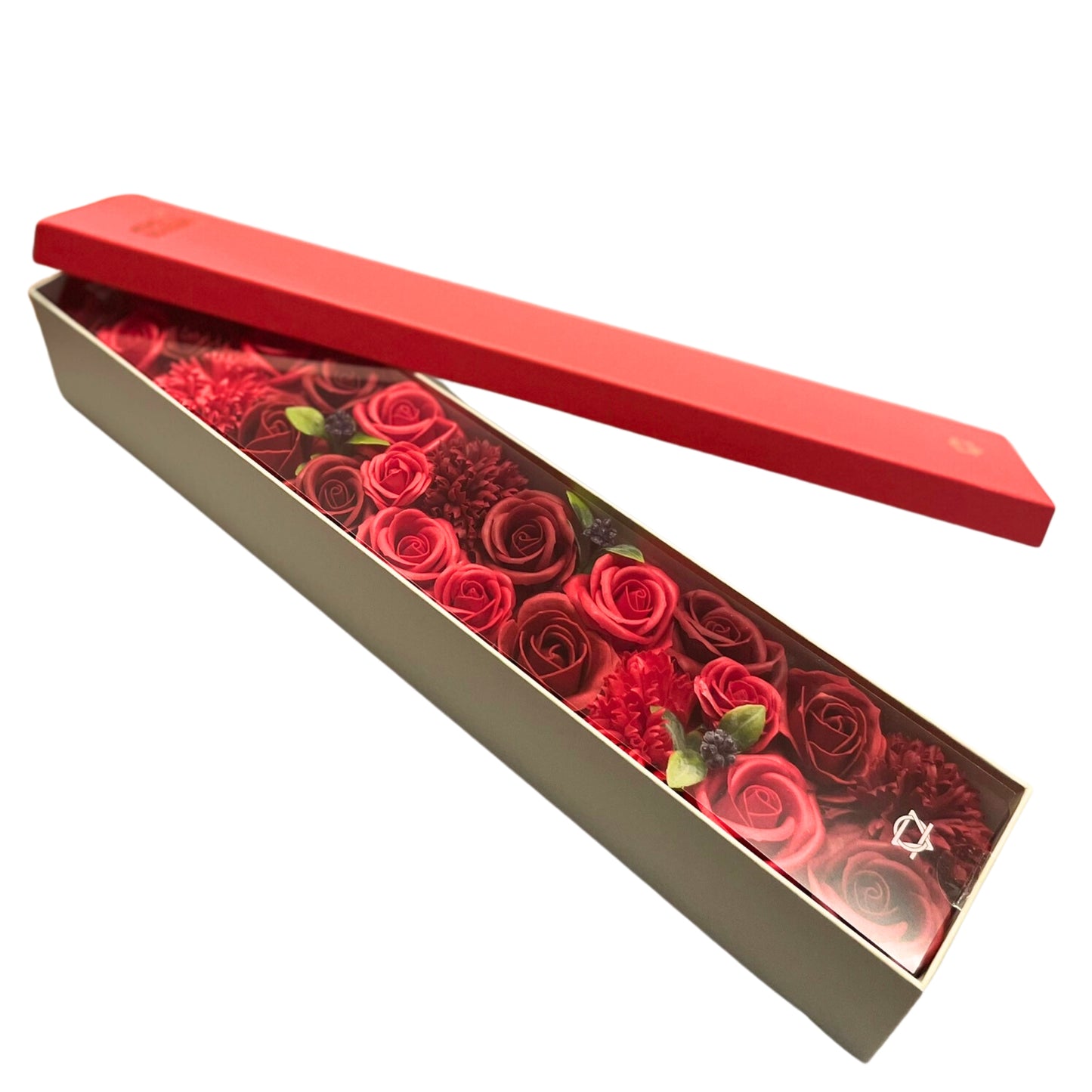 A long, rectangular box containing red soap roses and carnations is slightly opened, revealing its contents. The box is light beige, with a red lid. A logo is visible at the bottom right corner of the box.