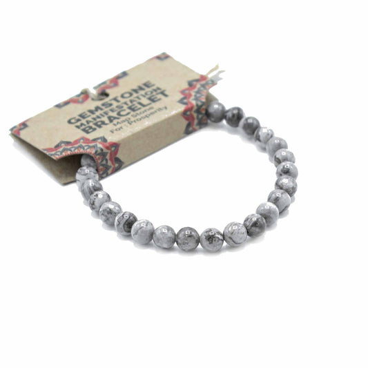 A grey gemstone bracelet, composed of round beads, rests on a white background; it is attached to a card that reads "GEMSTONE MANIFESTATION BRACELET Map Store For: Prosperity".