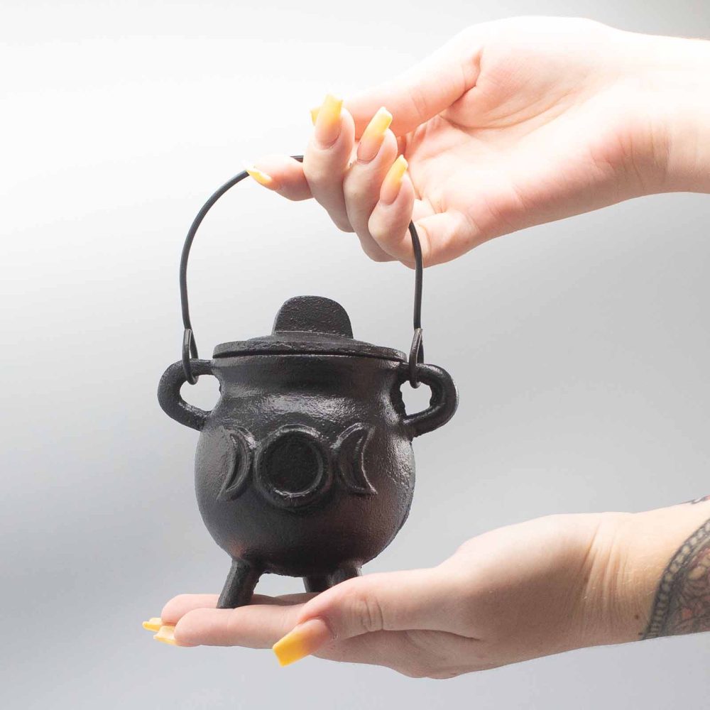 A small black cauldron with a lid is held between hands. It has a metal handle and features a triple moon design. The background is a neutral gray.