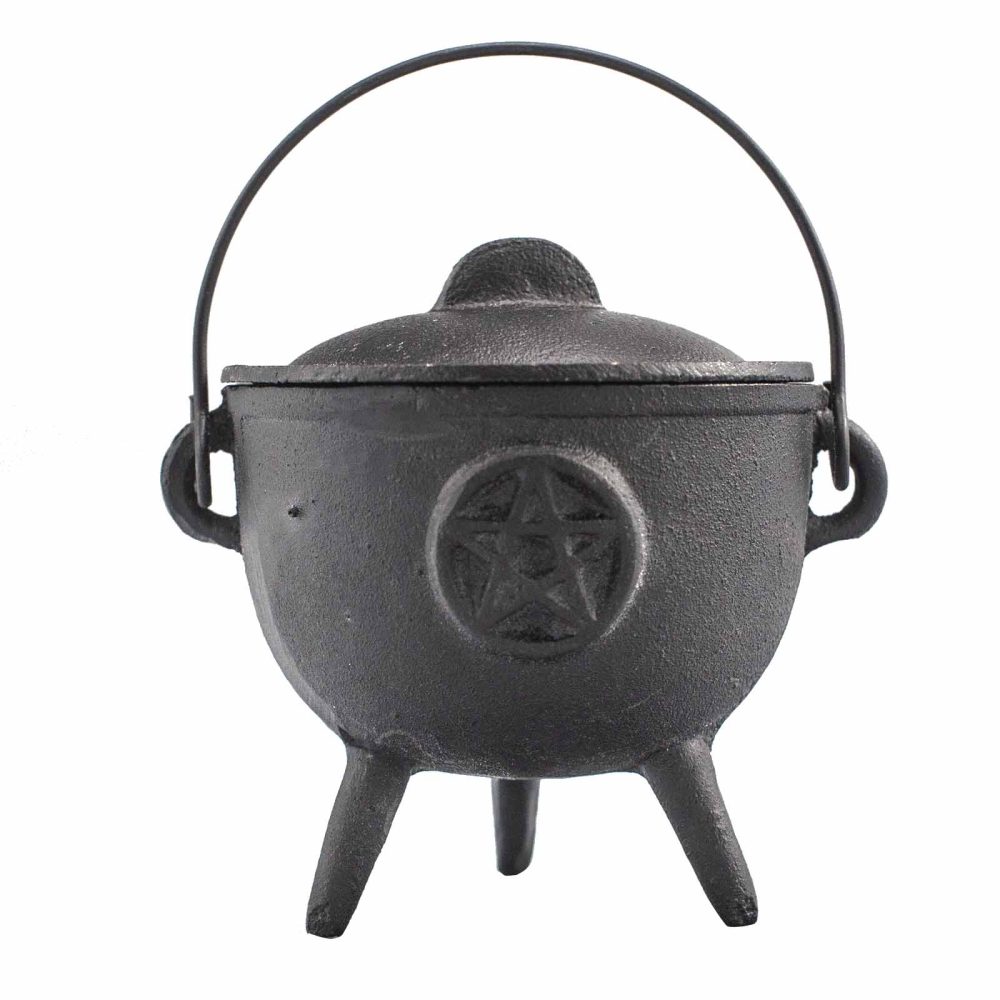 A black cast-iron cauldron with a lid and handle sits on three legs. A pentagram is embossed on its front. It rests in a plain, white setting.