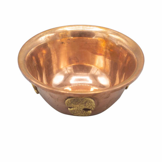 A polished copper bowl rests upright, reflecting light brilliantly. Embellished with small, decorative brass accents, it stands against a plain white background, emphasizing its warm metallic tones.