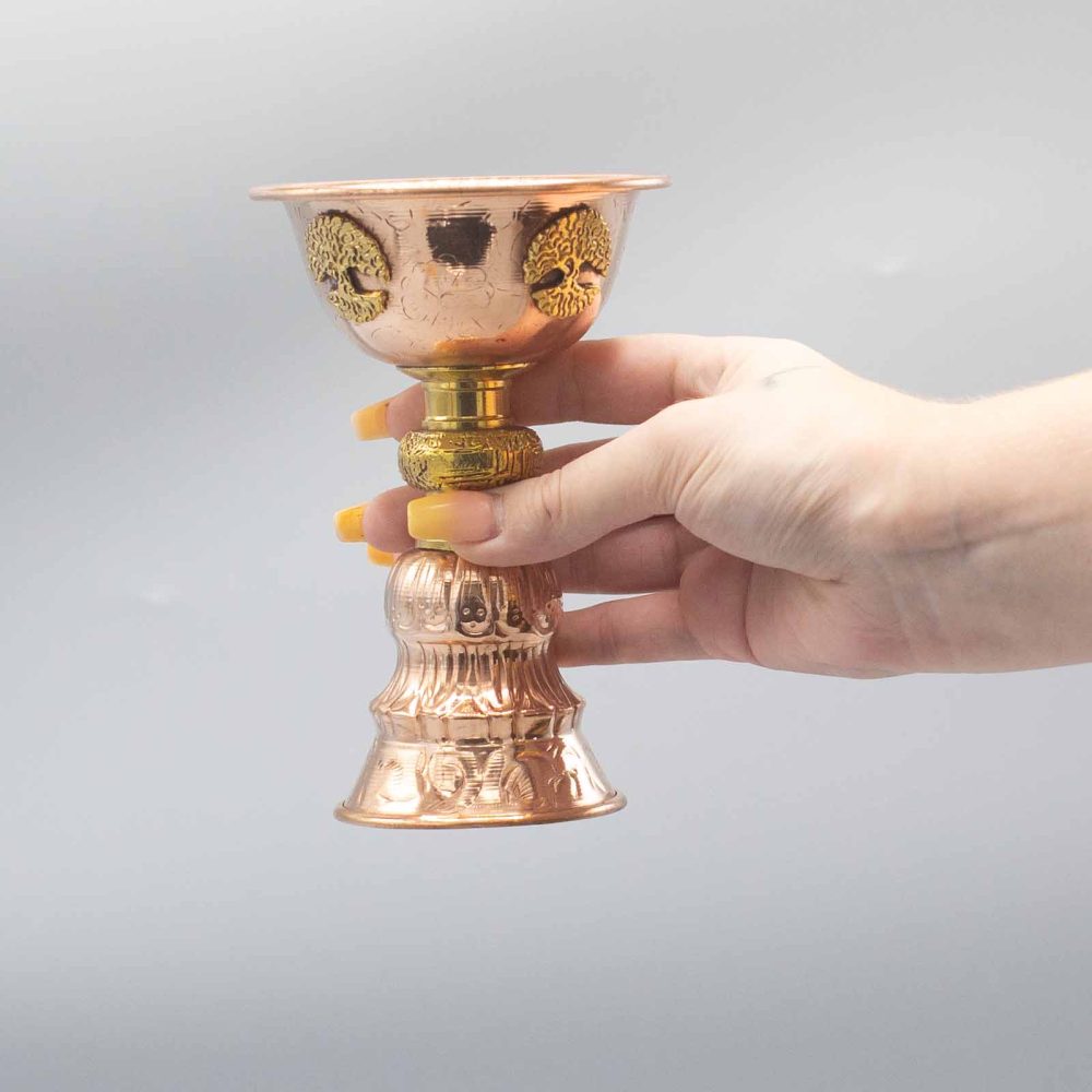 A detailed copper chalice is held in a hand against a plain background. The chalice features intricate engravings, with a wide base and decorated stem, showcasing craftsmanship.
