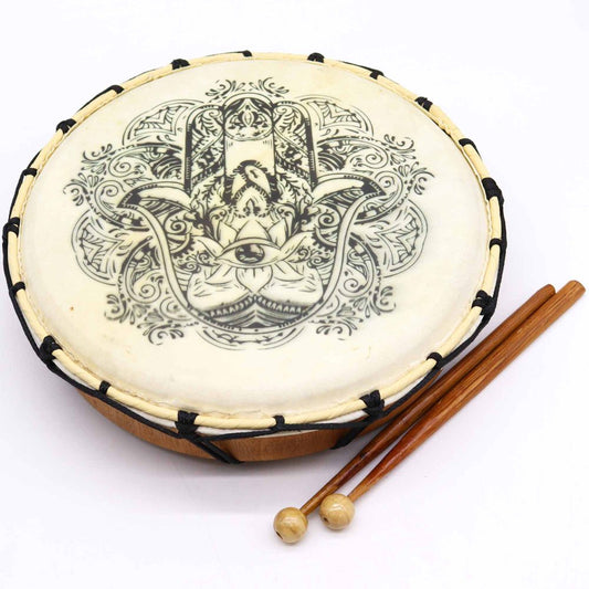 A decorated hand drum with intricate black patterns, featuring a detailed hamsa design, lies next to two wooden mallets on a white background.