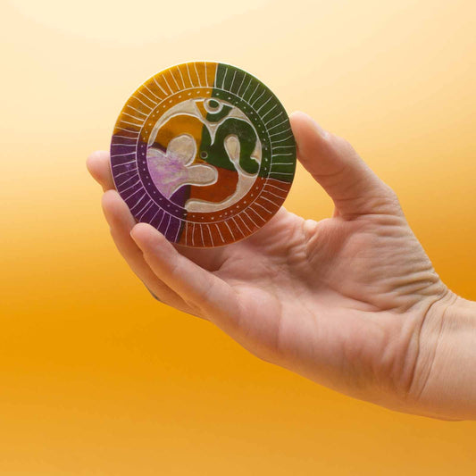 A hand holds a round, colorful disc with an abstract design featuring curved shapes and segmented colors. The background is a gradient of orange to yellow.