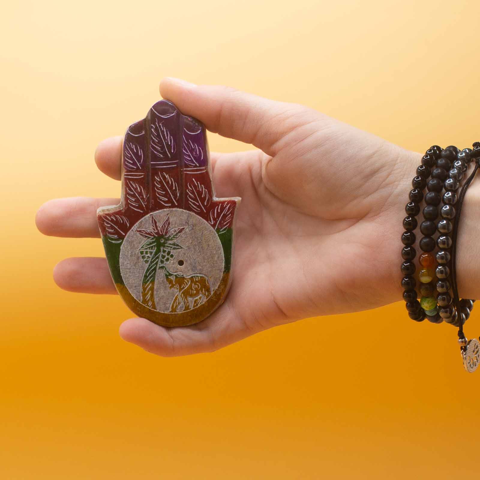 A hand holds a colorful Hamsa amulet with leaf patterns and an elephant beneath a tree, against a bright yellow background. The wrist is adorned with black beaded bracelets.