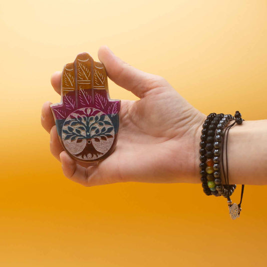 A hand holds a colorful, wooden hand-shaped object adorned with a tree design. The person wears black beaded bracelets. The background is a gradient of light to dark orange.
