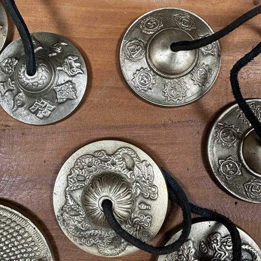 Bronze cymbals, intricately engraved with various circular symbols and patterns, are connected by black cords and placed on a wooden surface. The designs include dragons and floral motifs.