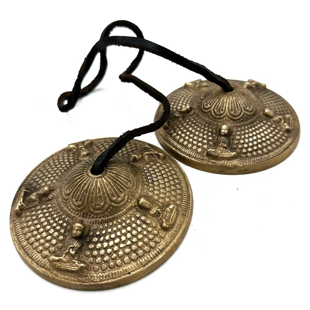 Two decorated brass cymbals with embossed patterns and figures are connected by a black string, resting on a white surface.
