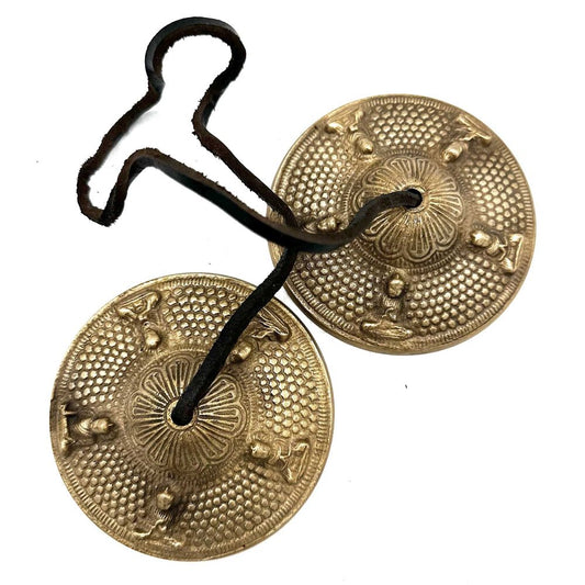 Two brass cymbals connected by a black leather cord; each cymbal features intricate engraved patterns and raised figures, lying against a white background.