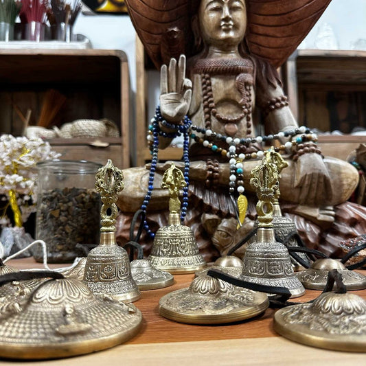 Brass bells are displayed on a wooden surface, surrounded by ornate cymbals. In the background, a wooden statue of a meditative figure with beaded necklaces creates a serene setting.