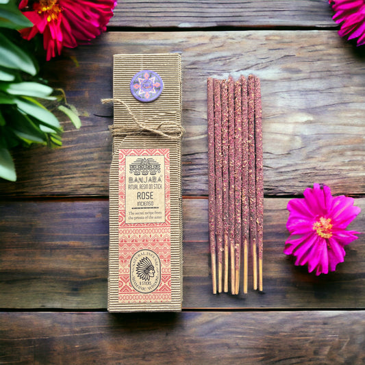 Incense sticks lie beside a textured box labeled "BANJARA RITUAL RESIN ON STICK ROSE incense," on a wooden surface, surrounded by vibrant pink flowers.