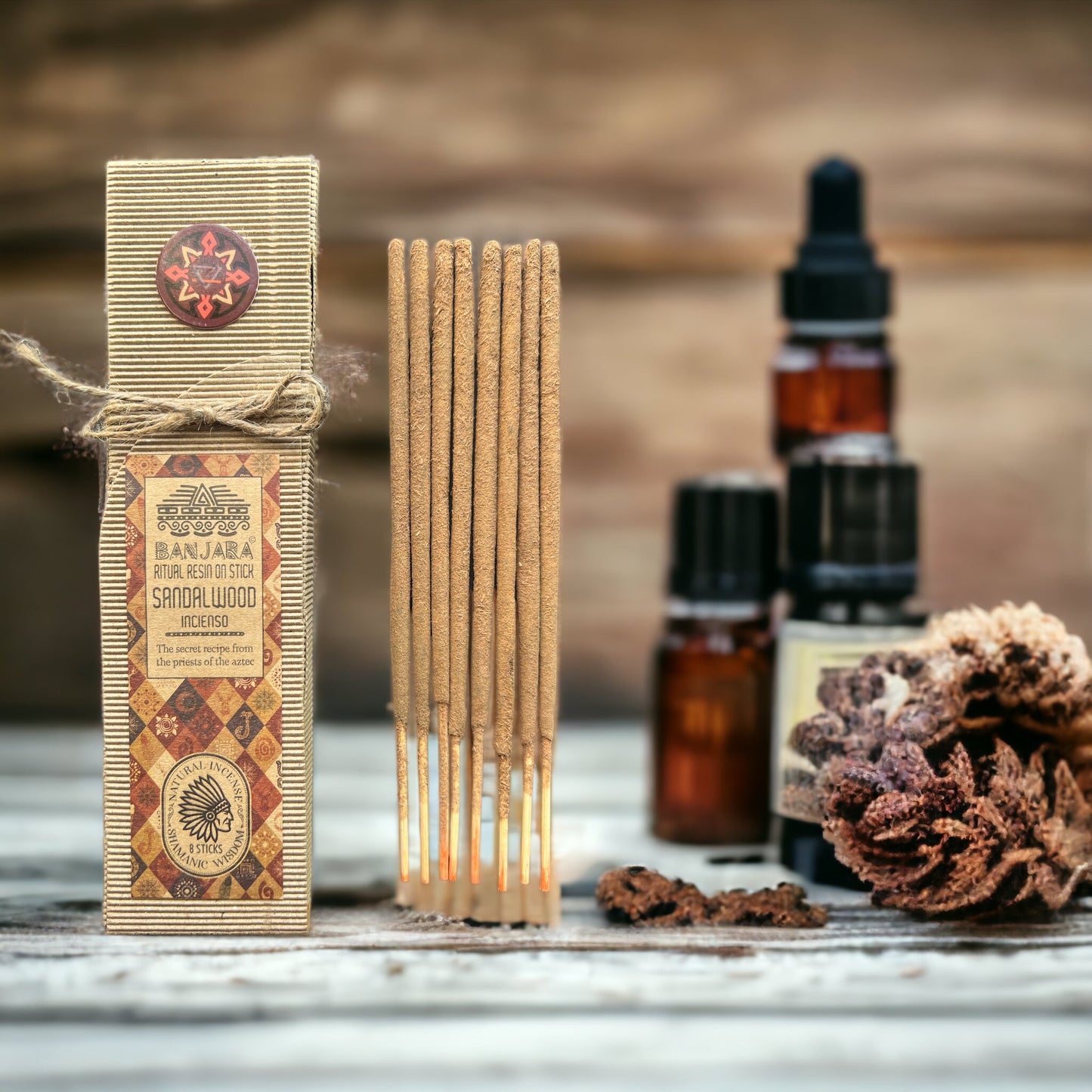 A box labeled "BANJARA Natural Resin on Stick Sandalwood Incense" stands beside burning incense sticks. Nearby, there are small amber bottles and dried botanicals on a rustic wooden surface.