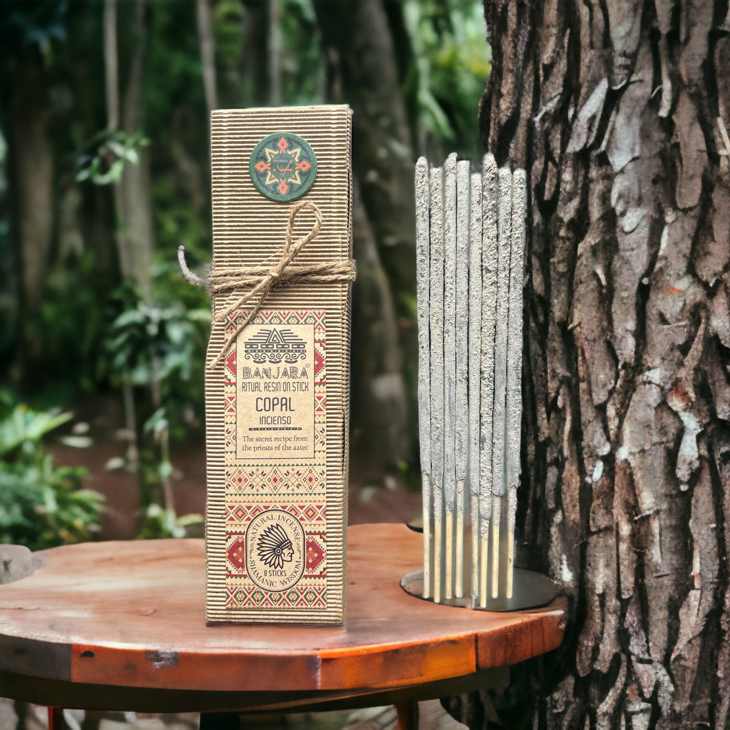 Package displaying "BANJARA RITUAL RESIN STICK COPAL incenso," wrapped with twine, stands on a wooden surface beside incense sticks near a tree. Text includes "The secret recipe from the priests of the Aztec."