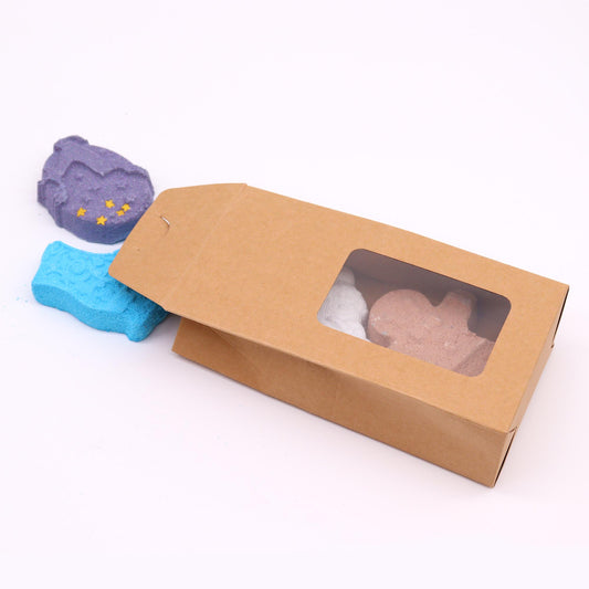 Colored bath bombs shaped like animals rest beside a rectangular brown box with a clear window on a white background.