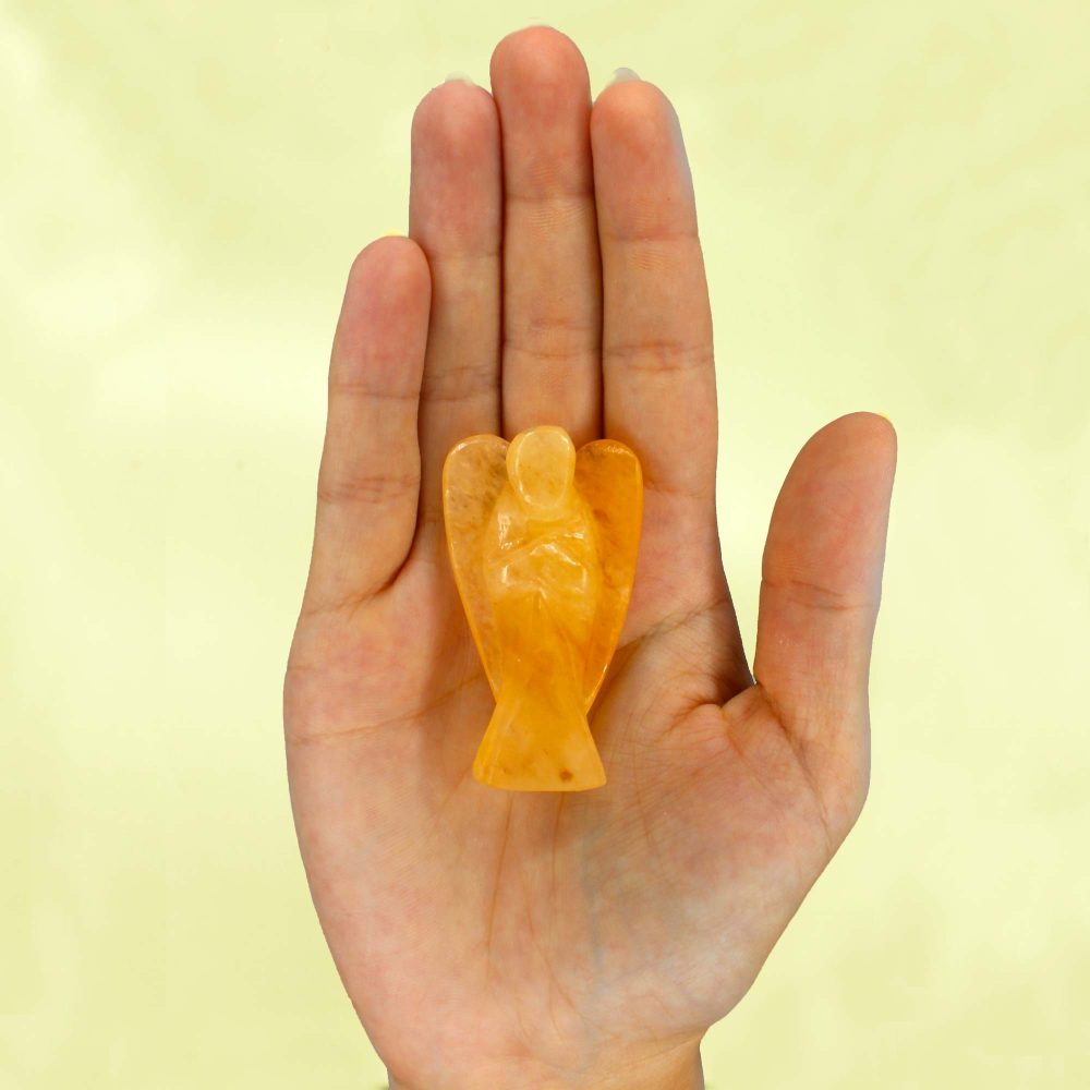 A small, light-orange stone angel rests in the center of an open human palm, set against a blurred light-yellow background.