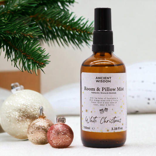 A brown glass bottle of room and pillow mist sits among Christmas ornaments and evergreen sprigs. The label reads, "ANCIENT WISDOM Room & Pillow Mist Habitación / Bruma de Almohada White Christmas 100ml e 3.38 fl oz".