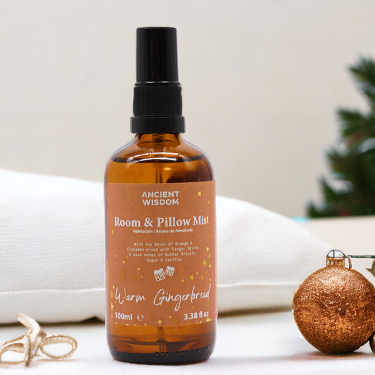 A brown glass bottle of room and pillow mist sits on a white surface near Christmas decorations. The label reads: ANCIENT WISDOM Room & Pillow Mist Habitación/Bruma de Almohada With Top Notes of Orange & Cinnamon mixed with Ginger Spices & base notes of Butter Biscuit, Sugar & Vanilla Warm Gingerbread 100ml e 3.38 fl oz