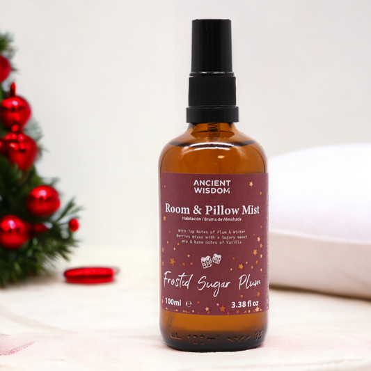 A room and pillow mist bottle sits on a white surface. It displays "ANCIENT WISDOM Room & Pillow Mist", "Frosted Sugar Plum", and volume information. A blurred Christmas-themed decoration is in the background.
