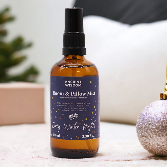 A room and pillow mist bottle sits on a surface. The bottle is amber glass with a black spray top. It is labeled "ANCIENT WISDOM Room & Pillow Mist," "Cosy Winter Nights," and includes scent descriptions.