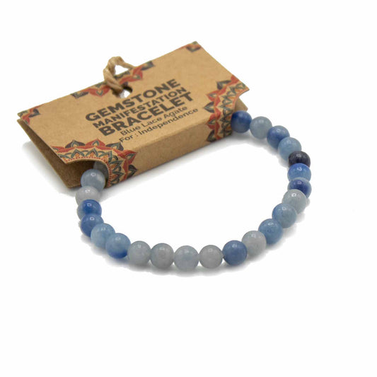 A gemstone bracelet, made of blue lace agate, rests on a white background; it is attached to a brown tag that reads "GEMSTONE MANIFESTATION BRACELET Blue Lace Agate For: Independence".