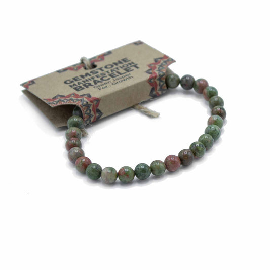 A gemstone bracelet, composed of green jasper beads, rests on a white background. Its accompanying card reads, "GEMSTONE MANIFESTATION BRACELET Green Jasper For Growth".