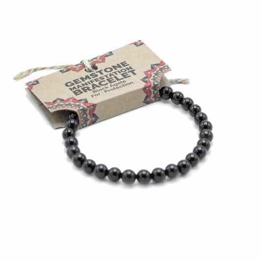 A black agate bracelet, composed of round beads, rests on a white background. It's accompanied by a kraft paper tag reading "GEMSTONE MANIFESTATION BRACELET Black Agate For: Protection".