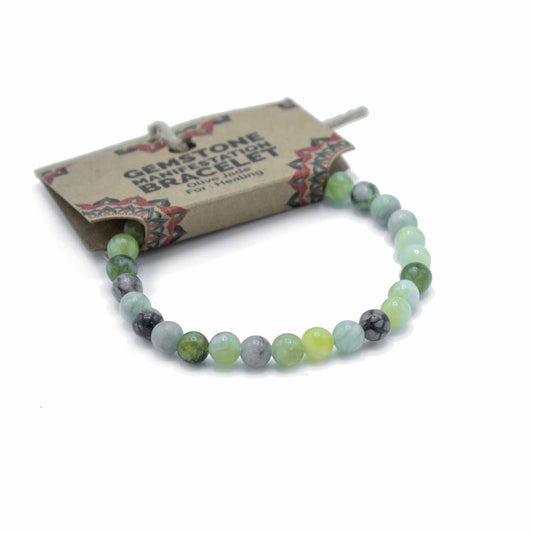 A gemstone bracelet, composed of various shades of green and grey beads, rests on a white background. The bracelet is attached to a brown card tag that reads: GEMSTONE MANIFESTATION BRACELET Olive Jade For Healing.