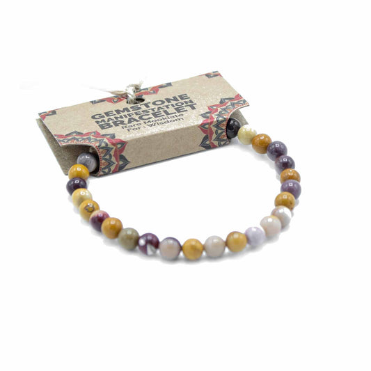 A gemstone bracelet, composed of multicolored beads, rests on a white background; it is packaged on a cardboard tag that reads "GEMSTONE MANIFESTATION BRACELET Rare Mookiate For Wisdom".