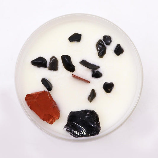 Round white candle in a clear glass holds a short wooden wick and scattered black and red polished stones set against a white background, photographed from above.