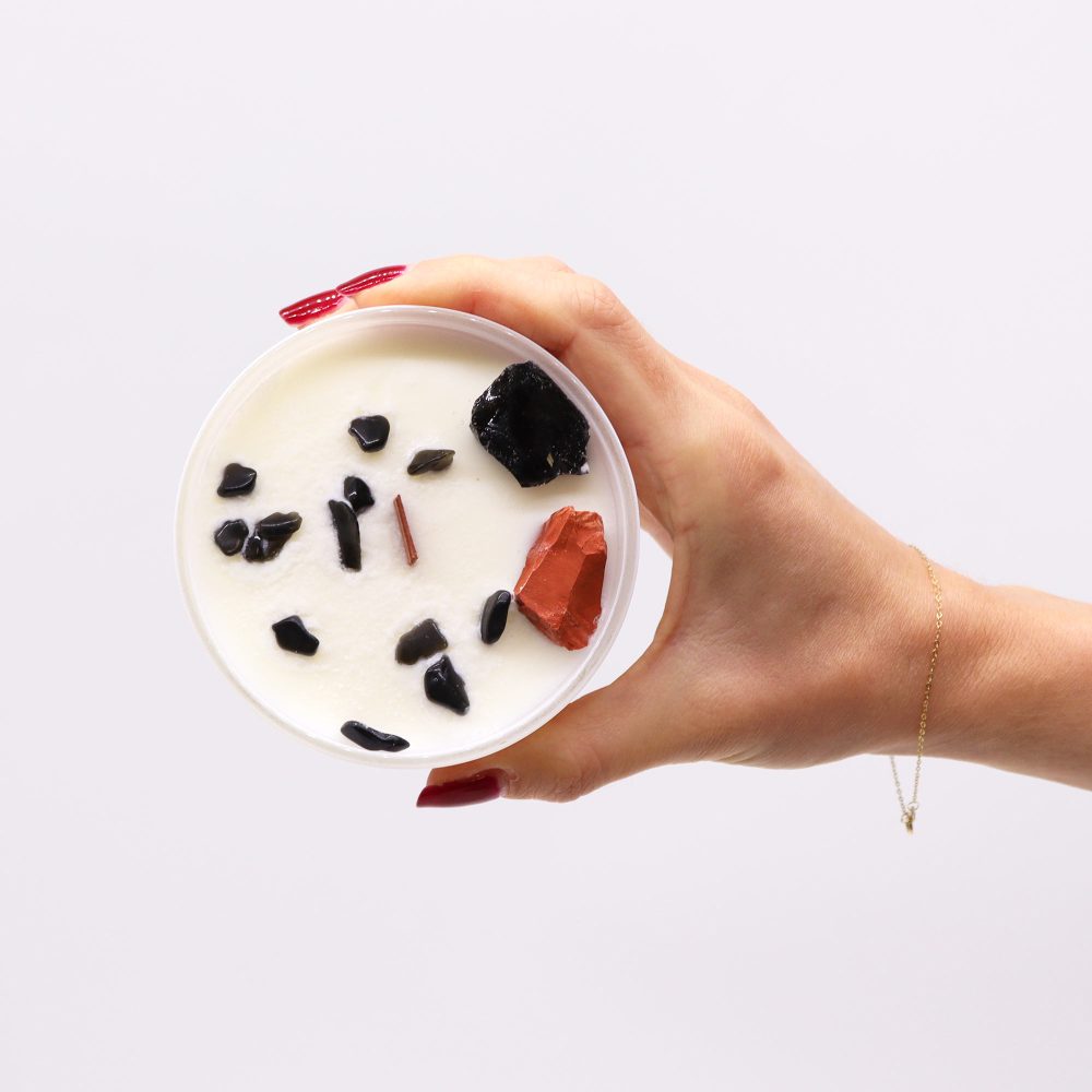 A hand with red nails holds a clear, round candle. The candle's wax surface has scattered black stones and one reddish-orange stone. The background is plain white.