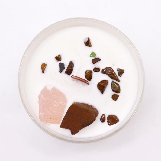 Round glass candle holds white wax sprinkled with small brown tumbled stones, a pink rose quartz chunk and a larger rust colored rock, photographed overhead on a white surface.