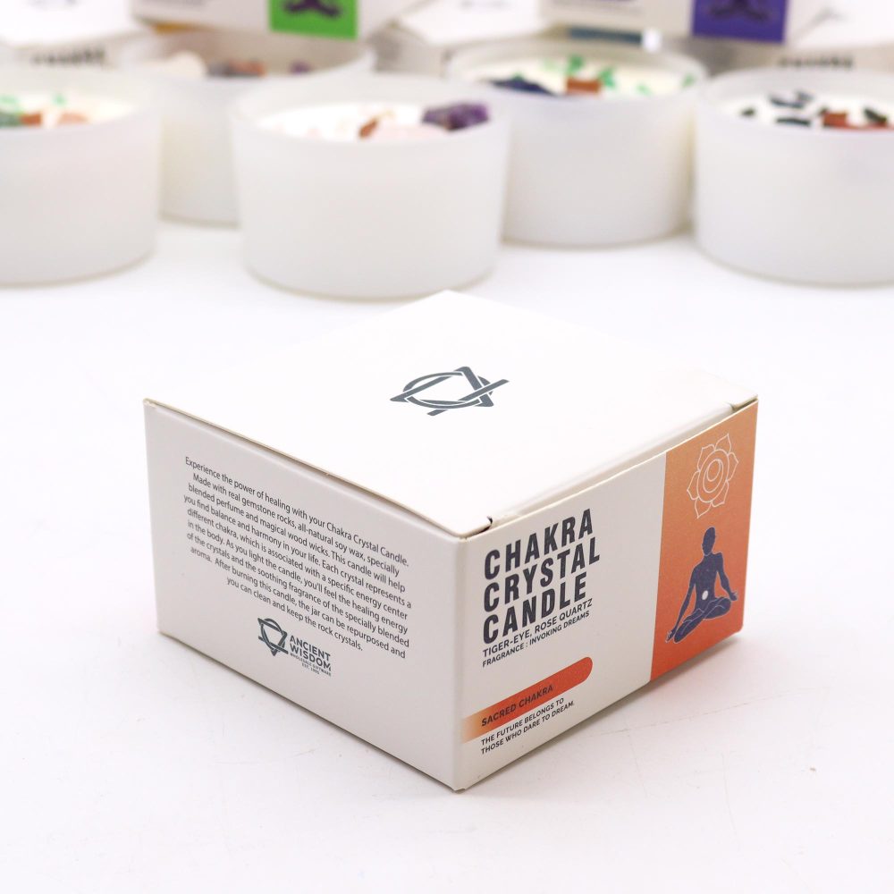A white box with "CHAKRA CRYSTAL CANDLE" printed on its side sits on a white surface, surrounded by blurry white candles topped with small stones. The box also says: "TIGER-EYE, ROSE QUARTZ, FRAGRANCE: INVOKING DREAMS, SACRED CHAKRA, THE FUTURE BELONGS TO THOSE WHO DARE TO DREAM."