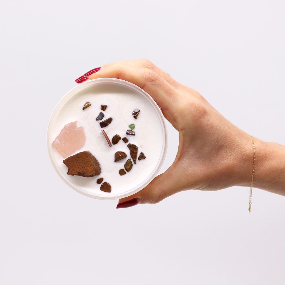 A hand with red nails holds a white candle filled with crystals and stones against a white background. The hand has a delicate gold chain bracelet.