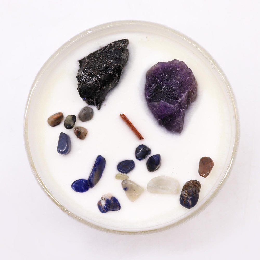 A round candle shows stones scattered in white wax, inside a clear jar. The stones include black, purple, and shades of blue, brown and tan. A small cinnamon stick also rests in the wax.