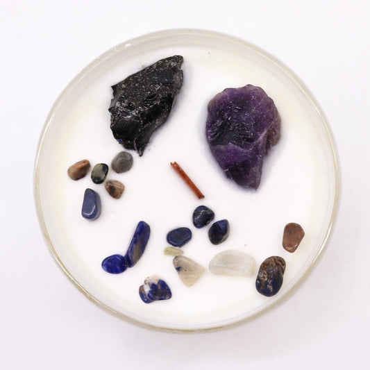 Round white candle in a glass dish holds scattered polished tumbled stones and two rough crystals on a plain white background.