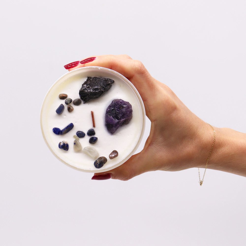 A white, round candle with several stones is held by a hand. The hand has red nail polish and wears a delicate gold bracelet. The background is white.