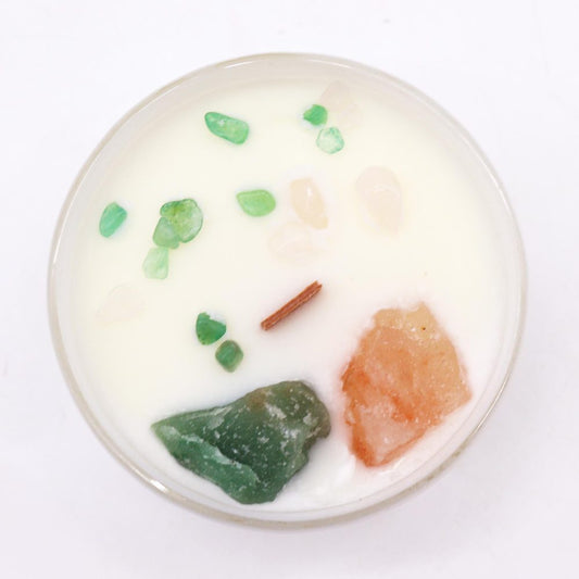 White wax candle holds small green and peach crystal chips and a short wooden wick, set inside a clear glass jar on a white surface viewed from above.
