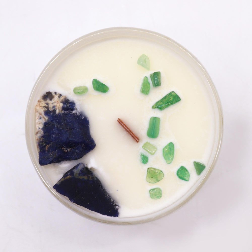 A candle, set in a beige jar, displays chunks of dark blue and small, green stones arranged on its top. A wooden wick is visible. The candle sits on a white surface.