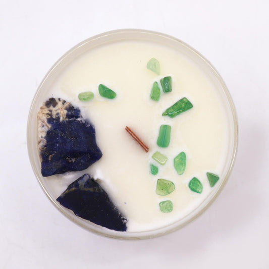 Round white wax candle sits in glass container with wooden wick, two irregular dark blue stones at left and scattered green glass chips at right on a white surface.