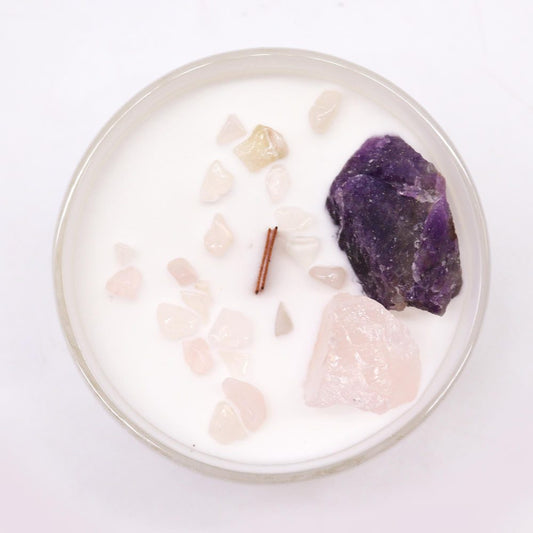 A round white candle holds a wooden wick and scattered pale pink chips plus a larger rose quartz and purple amethyst in a glass dish on a white surface.