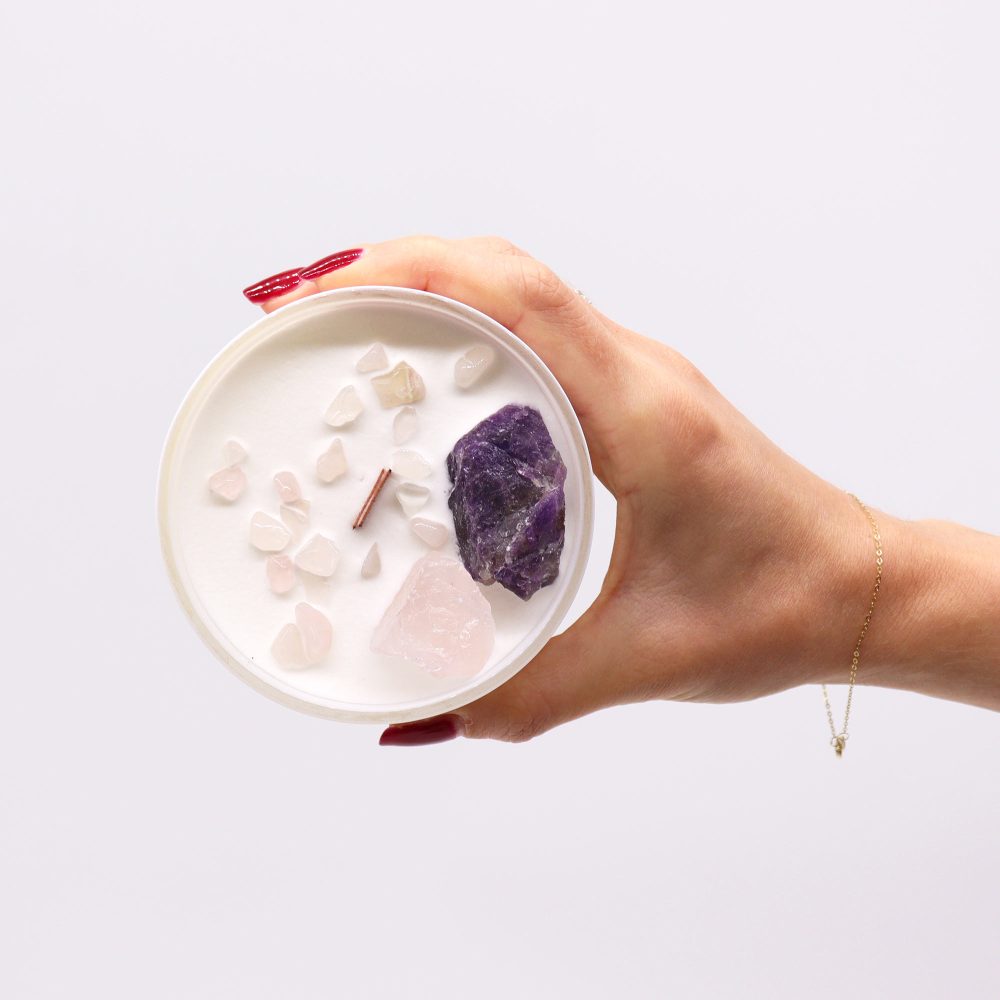 A manicured hand holds a white candle filled with white wax and various crystals and a copper wick centered on a white background.