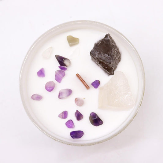 Round glass candle holds clustered amethyst chips, a large clear quartz, dark rough stone and a small cinnamon stick resting on white wax in a bright white tabletop setting.