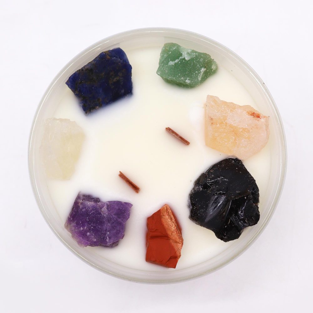 A candle sits on a white surface and contains chunks of multi-colored stones arranged around its edge, with two short, brown wicks in the center.