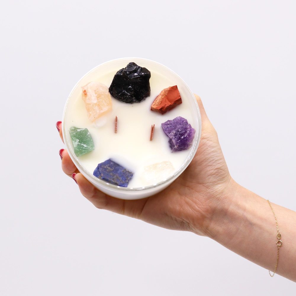 A candle, adorned with various crystals, is held by a manicured hand against a white background. The hand is wearing a delicate, gold bracelet.