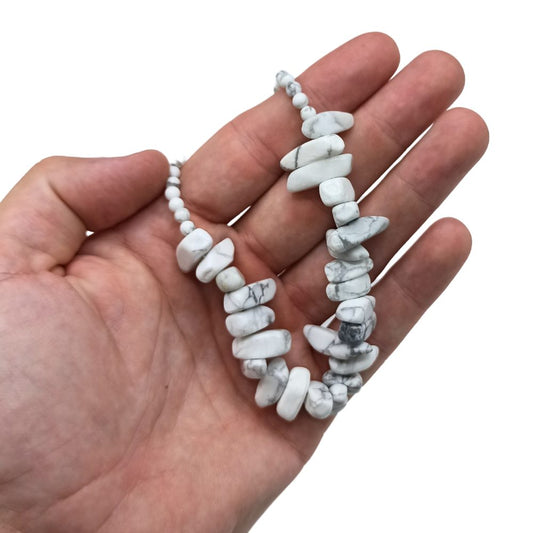 A white beaded necklace with grey markings is held in a light-skinned hand. The context is a plain white background.